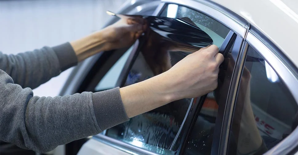Mãos a aplicar película escura no vidro lateral de um automóvel prateado, durante o processo de instalação ou verificação de películas nos vidros.