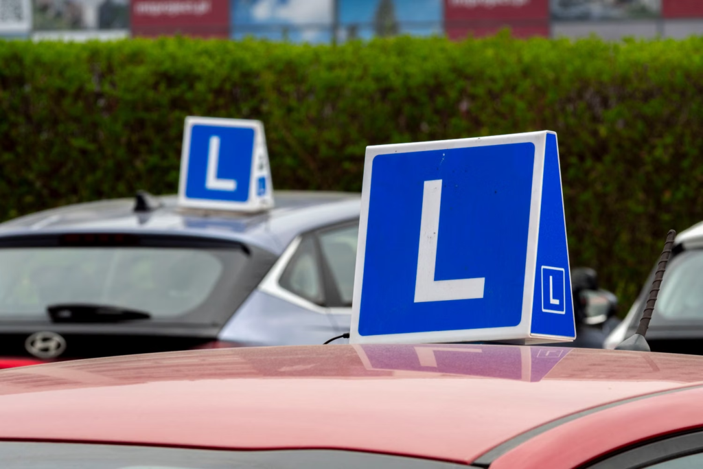 Um close-up dos tetos de dois carros de escola de condução estacionados, com destaque para a placa 'L' de instrução de condução azul e branca em primeiro plano, enquanto outra placa idêntica é vista no teto de um carro cinza borrado ao fundo